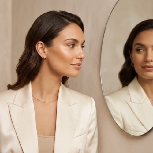 Captured with a high-resolution DSLR and an 85mm portrait lens, this image exemplifies professional editorial photography with a shallow depth of field. The subject, a refined woman, is bathed in soft, diffused natural light from a side angle, which delicately enhances the sharp contours of her jawline and cheekbones. Her complexion is flawless, featuring a subtle, healthy luminescence that catches the light naturally. She is dressed in an elegant, cream-colored satin blazer that drapes with a luxurious, shimmering texture, accented by minimalist gold-tone jewelry. The composition is balanced and introspective; she is captured in a quiet moment of self-reflection while looking into a circular mirror, which echoes her calm, poised expression. The background consists of a neutral, matte plaster wall, creating a clean, sophisticated, and minimalist aesthetic that allows the subject's natural grace to remain the focal point.
