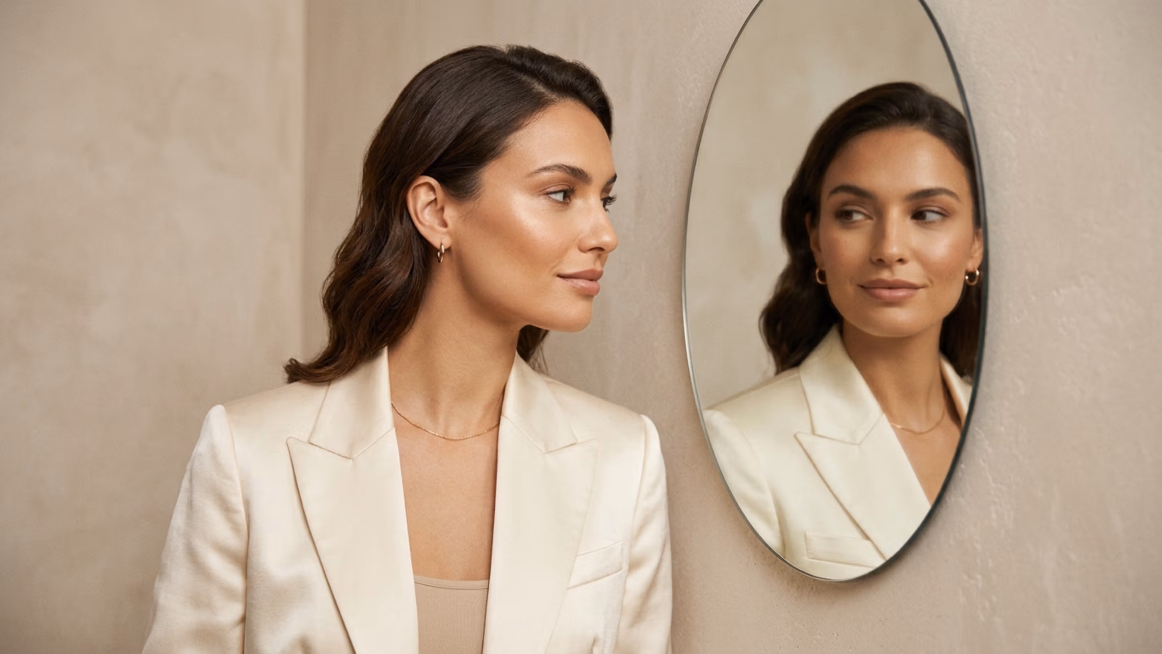 Captured with a high-resolution DSLR and an 85mm portrait lens, this image exemplifies professional editorial photography with a shallow depth of field. The subject, a refined woman, is bathed in soft, diffused natural light from a side angle, which delicately enhances the sharp contours of her jawline and cheekbones. Her complexion is flawless, featuring a subtle, healthy luminescence that catches the light naturally. She is dressed in an elegant, cream-colored satin blazer that drapes with a luxurious, shimmering texture, accented by minimalist gold-tone jewelry. The composition is balanced and introspective; she is captured in a quiet moment of self-reflection while looking into a circular mirror, which echoes her calm, poised expression. The background consists of a neutral, matte plaster wall, creating a clean, sophisticated, and minimalist aesthetic that allows the subject's natural grace to remain the focal point.