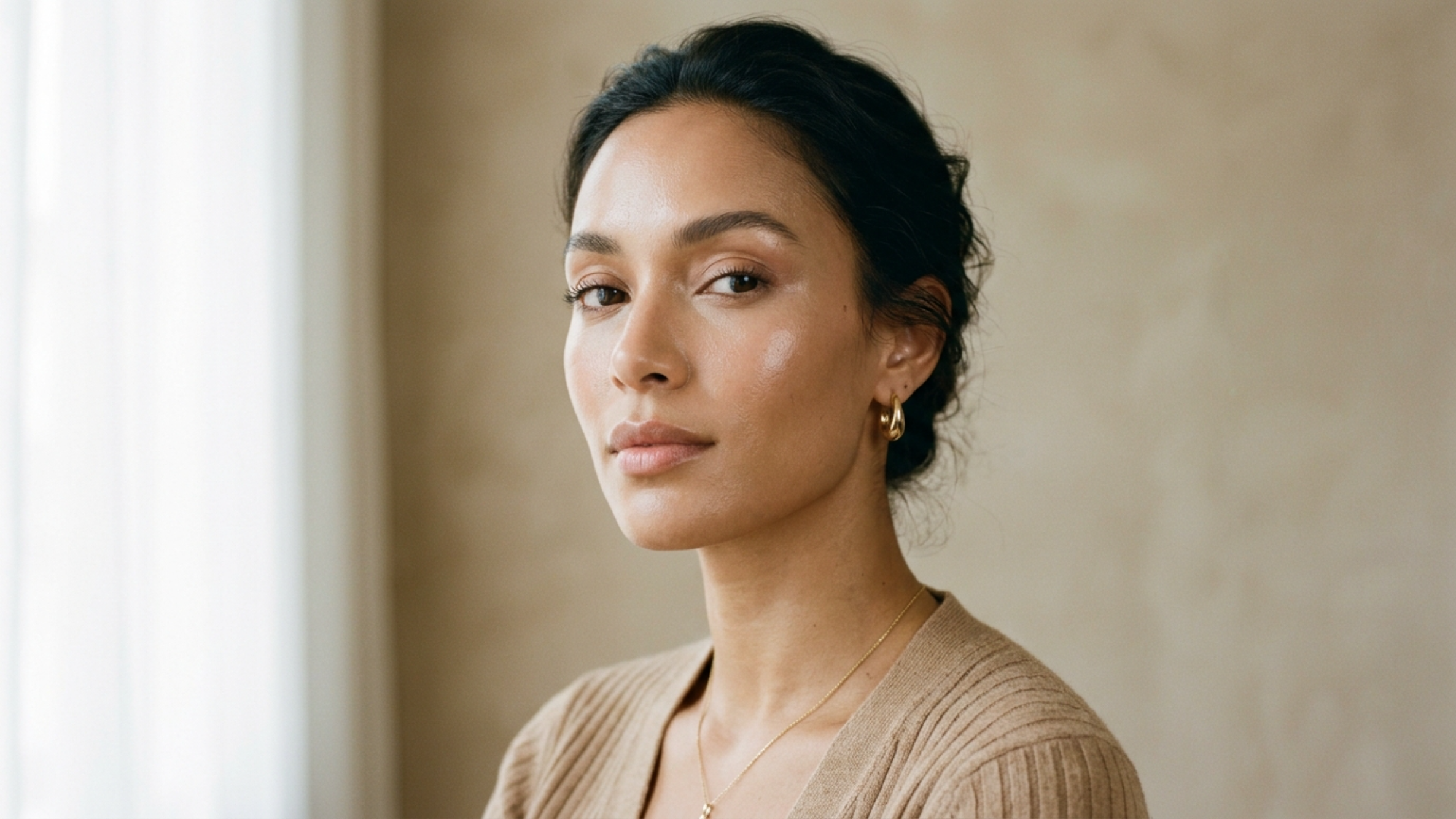 A high-resolution, professional editorial portrait of a woman with an olive complexion, captured with an 85mm prime lens for a shallow depth of field and exquisite detail. The lighting is soft and diffused, emanating from the left, which gently highlights the high points of her face and creates subtle, natural shadows that define her elegant bone structure. Her skin is depicted with a luminous, dewy quality, showing natural texture and subtle radiance. She is styled in a minimalist, beige ribbed knit sweater, complemented by understated gold hoop earrings and a delicate matching necklace. The composition is a sophisticated head-and-shoulders shot against a textured, neutral-toned wall, evoking a sense of modern, serene luxury. The overall aesthetic is clean, cinematic, and timeless.