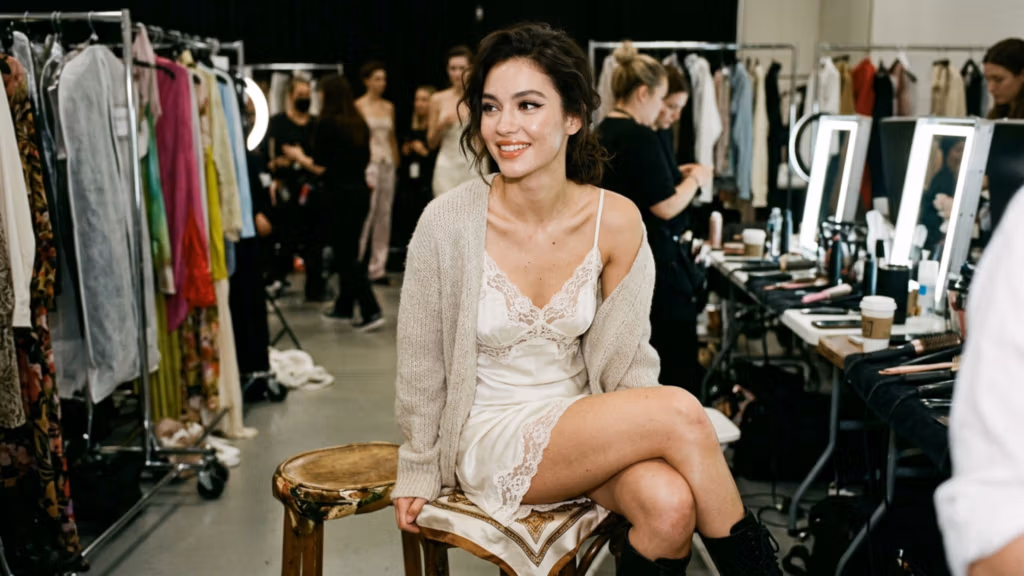 Ein professionelles Editorial-Foto fängt eine lächelnde junge Frau backstage bei einer Modenschau ein. Aufgenommen mit einer hochauflösenden DSLR und einem 50-mm-Objektiv (f/1.8) für eine geringe Schärfentiefe, die ihr ausdrucksstarkes Gesicht scharf fokussiert. Die Beleuchtung durch helle, sanft diffuse LED-Schminkspiegel erzeugt ein klares, helles Licht, das ihre Gesichtszüge wunderschön hervorhebt, die ebenmäßige Textur ihres Teints betont und ihrer Haut einen subtilen Schimmer verleiht. Die Frau mit feinen Gesichtszügen und einem fröhlichen, natürlichen Ausdruck sitzt im Schneidersitz auf einem Vintage-Hocker und strahlt eine anmutige und entspannte Haltung aus. Sie trägt ein zartes, cremefarbenes Satinkleid mit aufwendigen Spitzendetails, das unter einer kuscheligen, Oversize-Strickjacke getragen wird – eine gelungene Kombination aus sanftem Luxus und funktionalem Komfort. Die Szenerie ist ein geschäftiges Backstage-Ambiente mit Kleiderständern voller bunter Kleidung und Schminkplätzen, die eine authentische, industrielle Atmosphäre schaffen, die gleichermaßen lebendig und intim wirkt. Die Bildqualität ist gestochen scharf und klar und bewahrt eine elegante, ungezwungene redaktionelle Ästhetik.