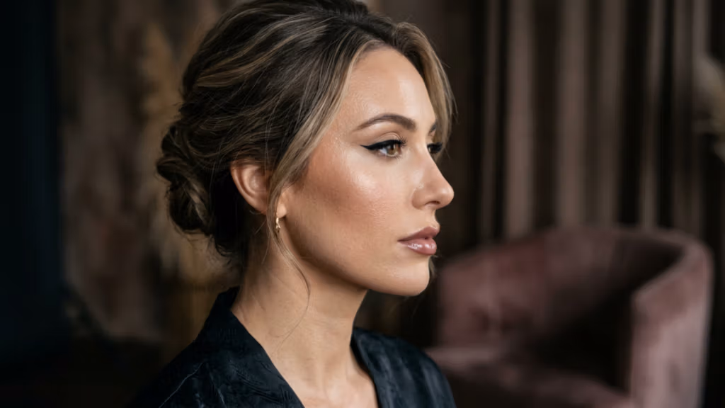 Captured with an 85mm portrait lens, this high-fidelity DSLR image showcases a sophisticated, professional aesthetic. The lighting is masterfully executed with a soft, directional side-light that emphasizes the subject's defined cheekbones and refined facial structure, creating a gentle gradient of shadow that adds depth and dimension. The female subject, positioned in a graceful profile, exhibits an elegant posture. Her complexion is captured with exquisite detail, revealing a healthy, dewy luminescence that glows under the studio lights, while her makeup is characterized by a sleek winged eyeliner and a glossy, natural lip. She is wearing a dark, subtly textured garment that suggests a luxurious silk or satin material. The background is a soft, out-of-focus interior space, featuring muted tones and a plush mauve chair, creating an intimate, high-end editorial atmosphere.