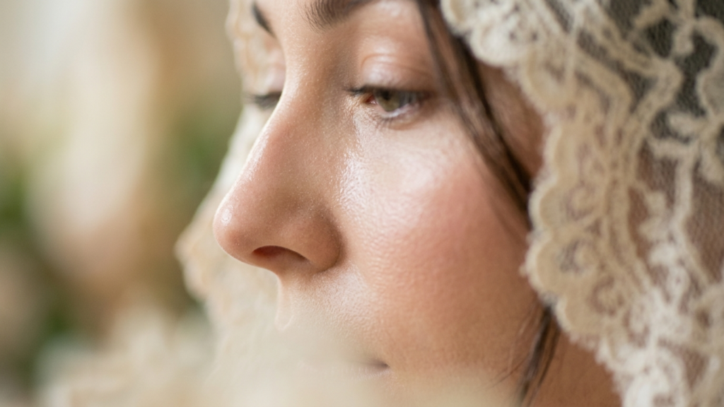 A high-end editorial close-up portrait captured with an 85mm macro lens, exhibiting the sharp, shallow depth-of-field characteristic of professional DSLR photography. The lighting is soft and directional, grazing the subject’s profile to highlight the natural, dewy texture of her skin, which glistens with subtle luminescence and microscopic moisture. The female subject, positioned in a pensive, downward gaze, is adorned in a delicate, vintage-style lace veil that frames her features with intricate, floral-patterned filigree. The focus is razor-sharp on the bridge of the nose and the cheekbone, while the background remains softly blurred into an ethereal, light-filled atmosphere, evoking a sense of timeless, romantic elegance. The color palette is warm and natural, emphasizing the organic interplay between the fine lace texture and the smooth, pore-detailed skin.