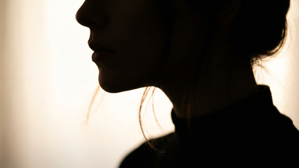 A high-end editorial close-up portrait captured in 4K resolution, utilizing a 85mm prime lens for a shallow depth of field that emphasizes the subject's profile. The lighting is masterfully executed with a high-contrast rim light technique, casting the subject into a dramatic silhouette against a soft, ethereal backlit backdrop. The composition focuses on the feminine contours of the jawline and neck, highlighted by a glowing, warm luminescence that traces the edges of the facial features. The subject wears a minimalist, dark-toned garment with a structured high neckline, suggesting a sleek, textile-rich fabric. The overall atmosphere is intimate and sophisticated, characterized by a refined chiaroscuro effect that accentuates the delicate stray strands of hair catching the backlight, creating a sense of quiet elegance and artistic tension.