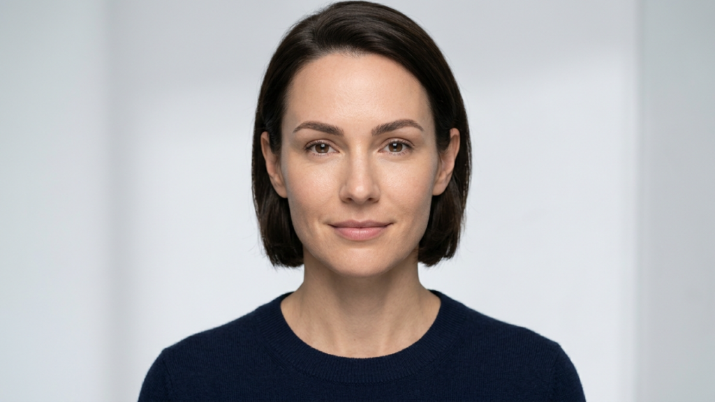 This high-resolution editorial portrait captures a woman with a serene expression, rendered with the precision of professional DSLR photography. Shot with a sharp, medium-telephoto lens—likely an 85mm—the image prioritizes subject clarity, isolating the subject from a clean, minimalist studio background. The lighting is soft and diffused, creating a flattering, even illumination across the face that minimizes harsh shadows while accentuating the natural, healthy skin texture and balanced facial features. The subject’s skin exhibits a natural, matte-like finish with lifelike micro-details, emphasizing a polished and professional aesthetic. She is wearing a minimalist, dark navy blue knit sweater with a fine-gauge texture, providing a subtle contrast against the high-key, neutral backdrop. The composition is a classic, centered head-and-shoulders portrait, conveying a sense of sophistication, accessibility, and calm confidence.