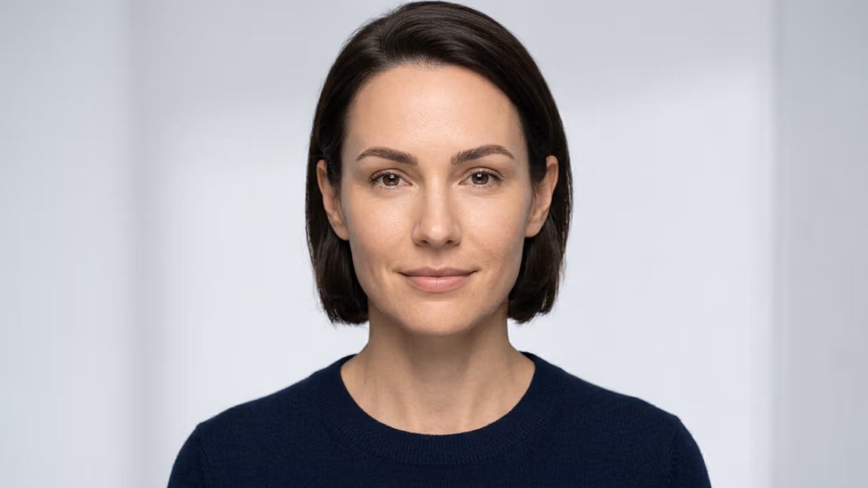 This high-resolution editorial portrait captures a woman with a serene expression, rendered with the precision of professional DSLR photography. Shot with a sharp, medium-telephoto lens—likely an 85mm—the image prioritizes subject clarity, isolating the subject from a clean, minimalist studio background. The lighting is soft and diffused, creating a flattering, even illumination across the face that minimizes harsh shadows while accentuating the natural, healthy skin texture and balanced facial features. The subject’s skin exhibits a natural, matte-like finish with lifelike micro-details, emphasizing a polished and professional aesthetic. She is wearing a minimalist, dark navy blue knit sweater with a fine-gauge texture, providing a subtle contrast against the high-key, neutral backdrop. The composition is a classic, centered head-and-shoulders portrait, conveying a sense of sophistication, accessibility, and calm confidence.