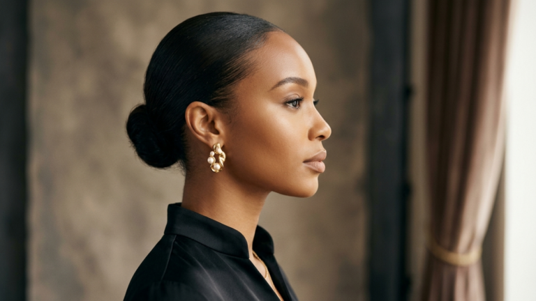 A high-end, professional editorial portrait captured with an 85mm prime lens, offering a shallow depth of field and pristine 4K resolution. The composition features a profile shot of a woman with an elegant, pulled-back hairstyle, highlighting her refined facial bone structure and smooth, luminous skin texture. The lighting is soft and diffused, creating a flattering, gentle illumination that emphasizes the contours of her jawline and features without harsh shadows. She is dressed in a minimalist, black silk garment, complemented by sophisticated, sculptural gold-and-pearl earrings that serve as a focal point. The background is a blurred, neutral-toned interior with soft drapery, evoking an atmosphere of understated luxury and timeless serenity.