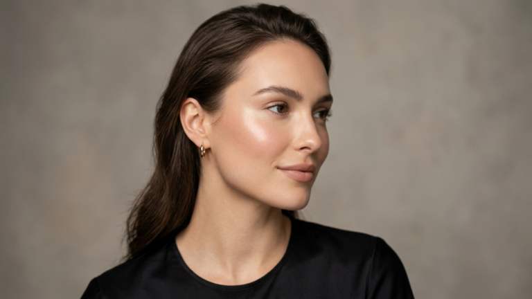 A professional high-end beauty editorial portrait captured with an 85mm prime lens, delivering exquisite DSLR-quality clarity and a shallow depth of field that isolates the subject. The lighting is soft and directional, emanating from the side to create subtle, flattering shadows that sculpt the subject's refined facial structure and highlight her elegant jawline. The subject is a woman with poised, classical features, exuding a serene and confident aura. Her skin displays a luminous, dewy texture with a natural, hydrated glow that catches the light beautifully on the cheekbones and bridge of the nose. She wears a minimalist black top crafted from high-quality, smooth fabric, complemented by simple, sophisticated gold hoop earrings. The composition is a clean, waist-up shot set against a soft-focus, neutral grey studio background, evoking an atmosphere of understated luxury and timeless elegance.