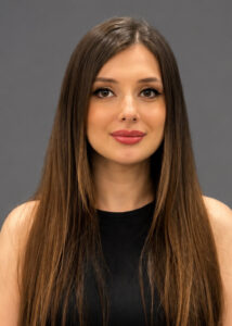 Ritratto professionale in studio di una giovane donna con lunghi capelli castani lisci, scattato con una fotocamera reflex digitale ad alta risoluzione. L'immagine è realizzata con un obiettivo da ritratto da 85 mm, che mette a fuoco i lineamenti del viso del soggetto mantenendo una ridotta profondità di campo. L'illuminazione è morbida e uniforme, tipica dei ritratti editoriali di alta gamma, minimizzando le ombre dure e creando una luminosità naturale sulla sua pelle liscia e luminosa. La modella ha un'espressione neutra ed elegante e indossa un semplice top nero senza maniche che conferisce un'estetica minimalista e moderna. Il soggetto è centrato nell'inquadratura su uno sfondo grigio opaco e pulito, che enfatizza un'atmosfera raffinata, professionale e senza tempo.