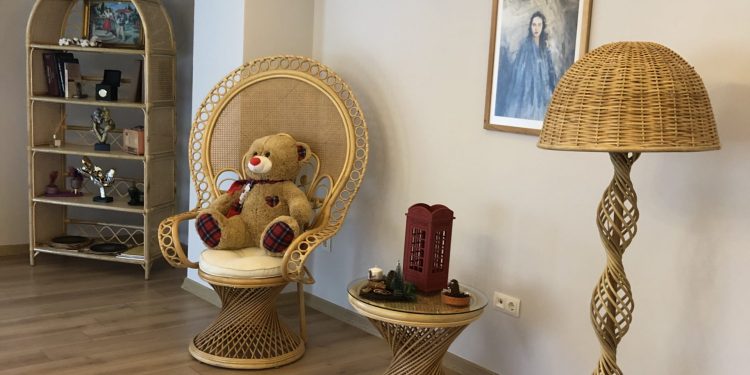 A teddy bear is sitting on a wicker chair in a room with a wooden floor. Next to the chair is a small wicker side table with a red telephone box decoration on it. There is also a tall wicker floor lamp with a spiral design and a framed picture hanging on the wall.