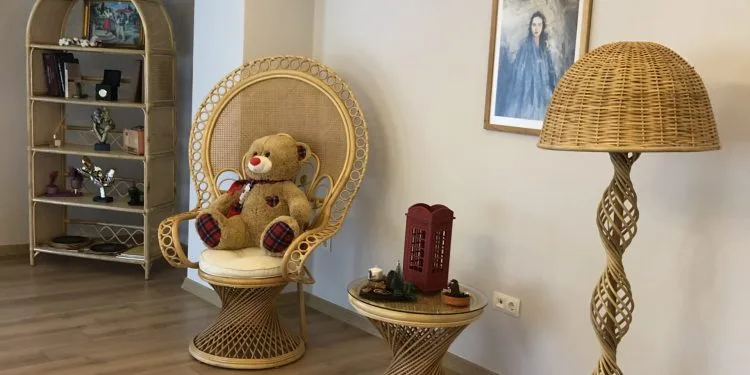 Un osito de peluche está sentado en una silla de mimbre en una habitación con suelo de madera. Junto a la silla hay una pequeña mesa auxiliar de mimbre con una decoración de cabina telefónica roja. También hay una lámpara de pie alta de mimbre con diseño en espiral y un cuadro enmarcado colgado en la pared.