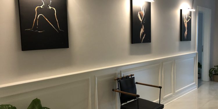 A hallway with three pieces of abstract art hanging on the wall, each illuminated by individual lights. The artwork features minimalist, monochromatic designs. A black chair with wooden armrests is positioned against the wall, and a large green plant is placed in the foreground. The hallway has white wainscoting and light-colored flooring.