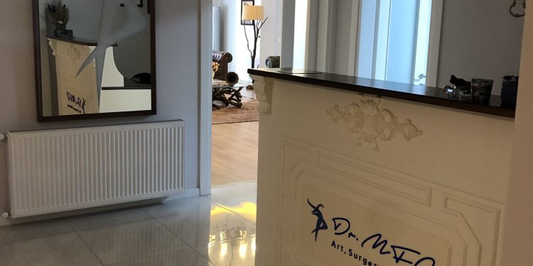 A modern reception area of Dr. MFO clinic featuring a white reception desk with 'Dr. MFO Art, Surgery & Beauty' written on it in blue. The area includes a large mirror, a radiator, and a well-lit hallway leading to another room with decorative elements.