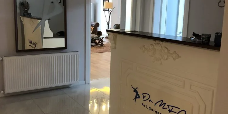 Una moderna recepción de la clínica Dr. MFO cuenta con un mostrador blanco con la inscripción "Dr. MFO Arte, Cirugía y Belleza" en azul. La zona incluye un gran espejo, un radiador y un pasillo bien iluminado que da acceso a otra sala con elementos decorativos.
