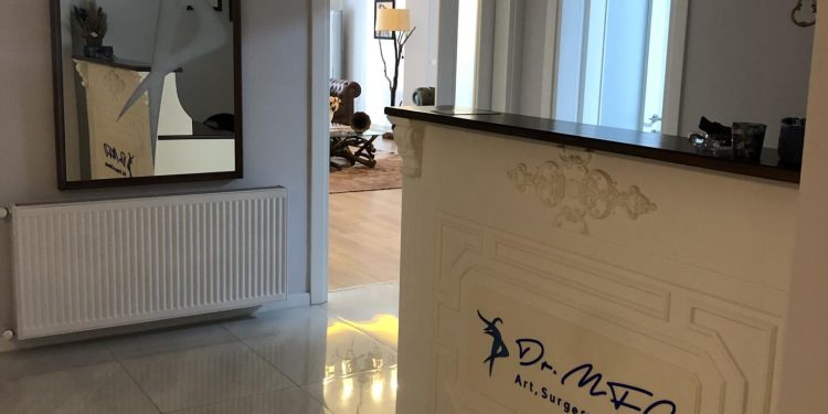 A modern reception area of Dr. MFO clinic featuring a white reception desk with 'Dr. MFO Art, Surgery & Beauty' written on it in blue. The area includes a large mirror, a radiator, and a well-lit hallway leading to another room with decorative elements.
