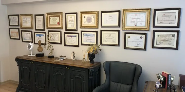 Imagen de una habitación con una pared adornada con múltiples certificados y diplomas enmarcados. Debajo de los certificados, hay un aparador de madera con diversos elementos decorativos, como un espejo, un jarrón con flores y una pequeña escultura. A la derecha del aparador, hay un sillón de aspecto cómodo, y a la izquierda, un escritorio con más elementos decorativos y un libro.