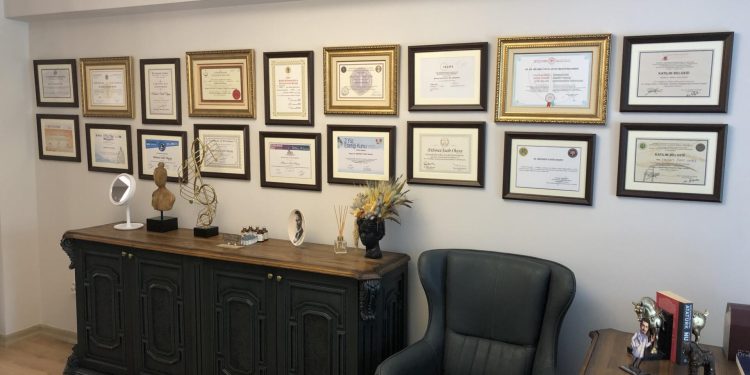 An image of a room with a wall adorned with multiple framed certificates and diplomas. Below the certificates, there is a wooden sideboard with various decorative items, including a mirror, a vase with flowers, and a small sculpture. To the right of the sideboard, there is a comfortable-looking armchair, and to the left, there is a desk with more decorative items and a book.