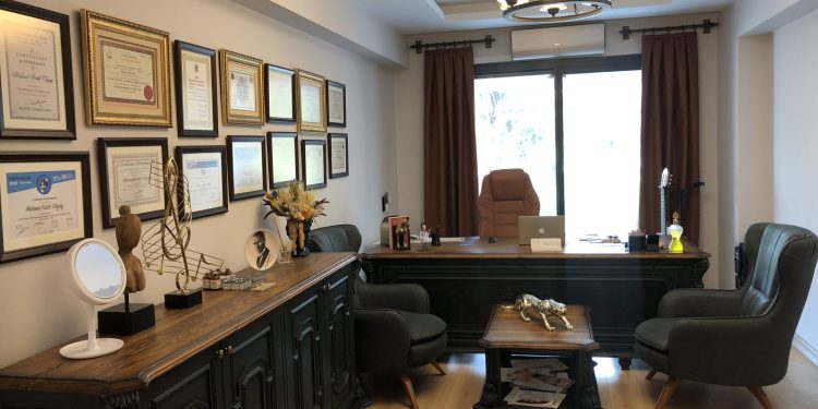 Uno spazio ufficio ben organizzato con una grande scrivania in legno attrezzata con cancelleria, un monitor per computer e oggetti decorativi. La scrivania è posizionata contro una parete con una grande finestra coperta da tende marroni. Certificati e premi incorniciati sono esposti sulla parete sopra la scrivania. Di fronte alla scrivania ci sono due sedie verdi e la stanza è caratterizzata da pavimenti in legno e un lampadario appeso al soffitto.