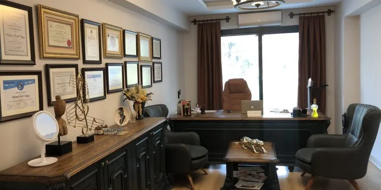 Una oficina bien organizada con un amplio escritorio de madera equipado con material de oficina, un monitor de computadora y artículos decorativos. El escritorio está ubicado contra una pared con una gran ventana cubierta por cortinas marrones. Certificados y premios enmarcados se exhiben en la pared sobre el escritorio. Frente al escritorio, hay dos sillas verdes, y la habitación cuenta con suelo de madera y una lámpara de araña que cuelga del techo.