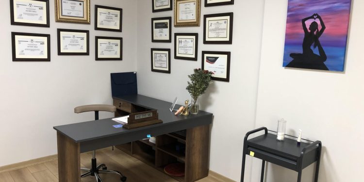 A home office setup featuring a desk with a computer, a chair, and a cart with exercise equipment. The wall is adorned with several framed certificates and a painting of a person in a yoga pose.