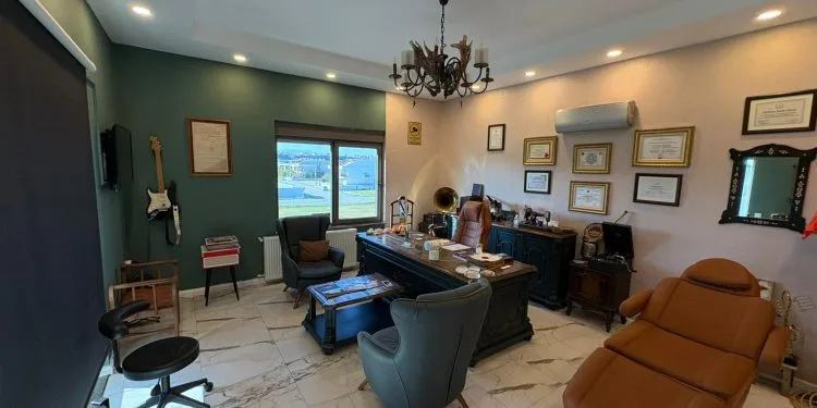 Un espacio de oficina profesional y decorado con estilo, que cuenta con un gran escritorio de madera, sillones de cuero, una camilla de exploración, una lámpara de araña ornamentada y diversos objetos decorativos en las paredes y estanterías.