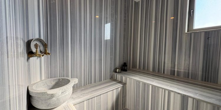 A modern Turkish bath interior featuring grey and white striped marble walls, a traditional marble basin with vintage-style brass faucets, and a sleek marble seating bench.