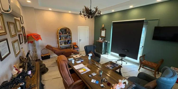 Un consultorio médico profesional y bien equipado, con escritorio de madera, sillas de cuero, maquetas médicas y una decoración elegante que incluye una lámpara de araña y una pared decorativa de color verde.