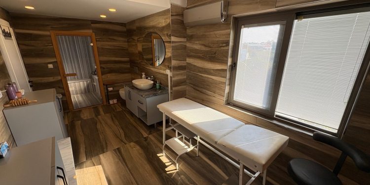 Modern medical or massage clinic room featuring a treatment table, a sink vanity, a mirror, and a small sauna enclosure with wood-patterned walls.