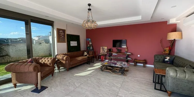 Una sala de espera de clínica moderna y acogedora, con sofás Chesterfield de cuero, una mesa de centro rústica de madera, una pared decorativa roja y una gran ventana con luz natural.
