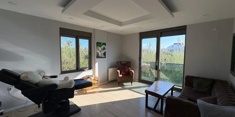Una moderna y luminosa sala de consulta con una silla de tratamiento con un osito de peluche, un cómodo sillón marrón, una mesa de centro de madera y un sofá marrón, con grandes ventanales con vistas a zonas verdes.